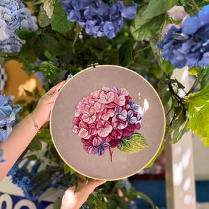 Puede incluir: Un bastidor de bordado circular de madera que contiene una tela gris con un dise&ntilde;o bordado a mano de una flor de hortensia. La flor presenta p&eacute;talos en tonos rosa, morado y azul con una hoja verde unida a un tallo marr&oacute;n.