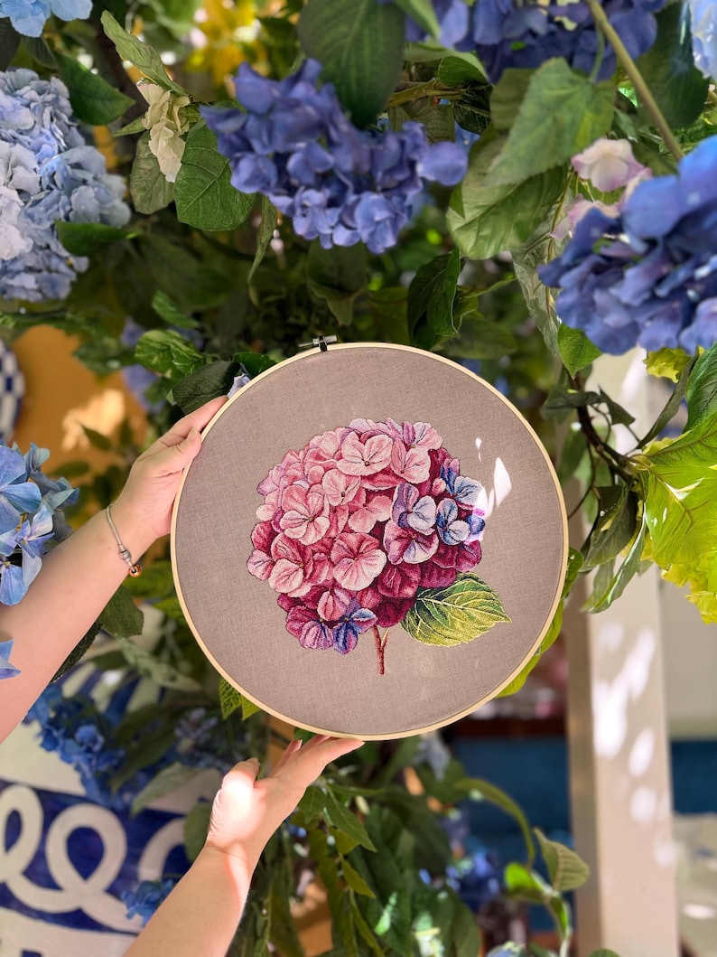 Puede incluir: Un bastidor de bordado circular de madera que contiene una tela gris con un dise&ntilde;o bordado a mano de una flor de hortensia. La flor presenta p&eacute;talos en tonos rosa, morado y azul con una hoja verde unida a un tallo marr&oacute;n.