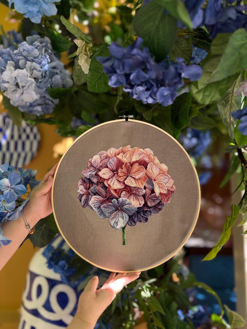 Puede incluir: Un bastidor de bordado circular de madera que contiene un panel de tela con un dise&ntilde;o detallado de una flor de hortensia. La flor presenta un degradado de p&eacute;talos rosas, melocot&oacute;n y morados, con un peque&ntilde;o tallo verde sobre un fondo gris.