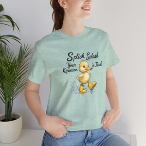 Op de afbeelding: Een lichtgroen t-shirt met een cartoon eend die gele regenlaarzen draagt. De tekst op het shirt luidt "Splish Splash Your Opinion is Trash".