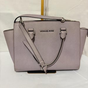 May include: A light purple Michael Kors handbag with a top handle and a detachable shoulder strap. The bag has silver-tone hardware and the brand name is displayed on the front. The bag has a rectangular shape with a structured design.
