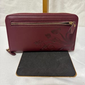 May include: A burgundy wallet with a gold zipper and a floral design. The wallet is rectangular and has a zipper closure. The floral design is embossed on the front of the wallet.