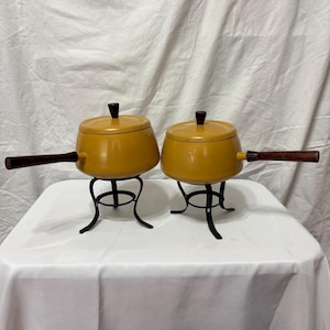 May include: Two mustard-yellow fondue pots with lids and wooden handles. Each pot sits on a black metal stand. The pots are on a white surface against a white backdrop.
