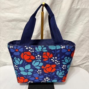May include: A navy blue tote bag with a floral pattern in red, light blue, and white. The bag has navy blue handles and a navy blue interior. The bag is sitting on a white surface.