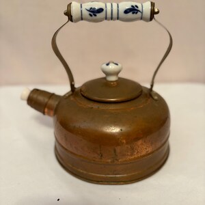 May include: A copper tea kettle featuring a rounded body, a short spout, and a lid topped with a white ceramic knob. The arched metal handle is fitted with a white ceramic grip decorated with blue leaf patterns, providing a contrast to the metal.