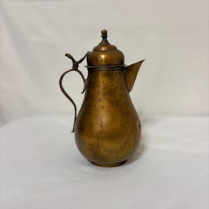May include: A vintage copper pitcher with a lid and a curved handle. The pitcher has a spout for pouring and a decorative finial on top. The copper has a warm, aged patina, suggesting it is an antique or a well-loved piece.