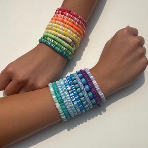 May include: Two wrists are shown, each wearing multiple stacks of flat disc bracelets. One wrist features a rainbow of colours, the other displays shades of blue, teal, and purple. The bracelets appear to be made of small, flat beads.
