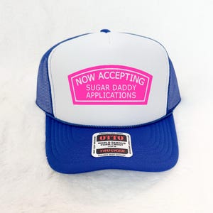 May include: A blue and white trucker hat with a pink rectangular patch that reads "NOW ACCEPTING SUGAR DADDY APPLICATIONS." The hat has a mesh back and a curved brim. The hat is made of foam fabric.