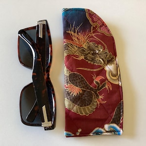May include: Tortoiseshell sunglasses with silver detailing beside a burgundy quilted glasses case. The case features a gold and black dragon design with red and blue accents. The sunglasses have dark lenses.