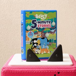 May include: A blue hardcover book titled Bluey 5-Minute Stories featuring a colorful illustration of the Bluey characters stacked on top of each other in a park setting with trees and hanging lanterns, held upright by two black triangular bookends.
