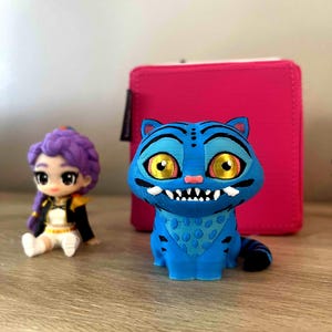 May include: A blue 3D-printed cat figurine with yellow eyes and a white-toothed grin, a small doll with purple hair, and a pink case sit on a wooden surface. The cat has a blue bandana and black stripes. The doll is wearing a black jacket.