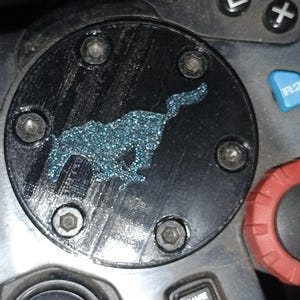 May include: Black circular component with a blue glitter silhouette of a running animal, possibly a horse. The component is secured with six silver screws and is part of a gaming controller.