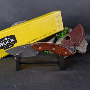 May include: A Buck Knives brand folding knife with a brown wooden handle and a black blade. The knife is displayed on a black stand with a yellow box behind it. The box has the text "Buck Knives" and "The measure of a knife since 1902".