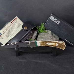 Könnte beinhalten: Ein Buck Knives Taschenmesser mit einem Hirschhorn-Griff, grünen Akzenten und einer gezahnten Klinge. Das Messer wird auf einem Ständer mit einer Lederscheide und einem Echtheitszertifikat präsentiert.