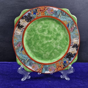 May include: A square-shaped decorative plate with a scalloped edge. The plate features a vibrant green center, a red inner border, and a detailed outer border with floral and landscape designs in various colors. The plate is displayed on a clear stand.