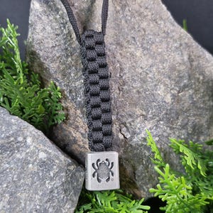 May include: A black paracord necklace with a silver charm featuring a spider design. The necklace is displayed against a background of rocks and greenery.