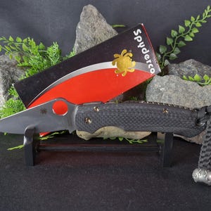 May include: A black Spyderco pocket knife with a black carbon fibre handle and a silver blade. The knife is on a black stand and is next to a red and black box with the Spyderco logo.