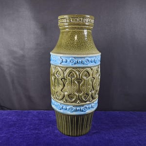May include: A tall, ceramic vase with a textured, olive green body and a light blue band. The vase features a repeating geometric pattern and a flared base. The top rim has a decorative band. The vase is a decorative home decor item.