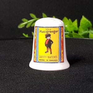 May include: White ceramic thimble with a colorful illustration of a boy wearing a red hat and black clothes. The illustration is surrounded by a yellow border with the text "Wee MacGregor Safety Matches Made in Sweden".