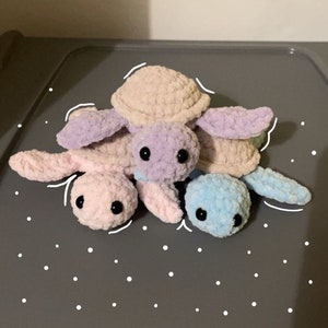 Op de afbeelding: Vier gehaakte schildpad knuffels in verschillende kleuren: roze, paars, lichtblauw en beige. De schildpadden zijn op elkaar gestapeld, met de beige schildpad bovenop.