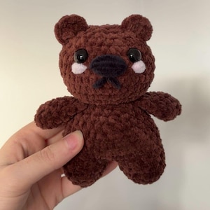 May include: A brown, handmade teddy bear with a soft, fluffy texture. The bear features black eyes, a black nose, and small pink accents on its cheeks. A cuddly, plush toy.