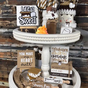 Peut inclure: Un présentoir blanc à deux niveaux avec des décorations sur le thème des s'mores. L'étage supérieur comporte une pancarte avec l'inscription « Life is Sweet », un pot de chocolat et une tasse de guimauve. L'étage inférieur comporte une pancarte avec l'inscription « You Make My Heart Melt » et un rouleau à pâtisserie avec l'inscription « Smores Bar ».