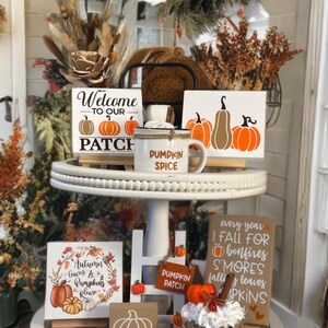 May include: A display of autumn-themed decorations on a tiered tray. Features signs with pumpkin designs and phrases such as "Welcome to our Patch" and "Pumpkin Spice." A mug, decorative pumpkins, and a faux dessert are also present.