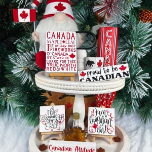 May include: A display of Canadian-themed ornaments on a tiered stand. Features a gnome, small flags, and signs with text such as "Canada" and "Proud to be Canadian." The colour scheme is red, white, and black.