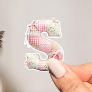 May include: A fabric letter "S" in a patchwork design, featuring pink, white, and red gingham and polka dot patterns. The letter is adorned with small pink bows and white lace accents, creating a decorative and handcrafted appearance.