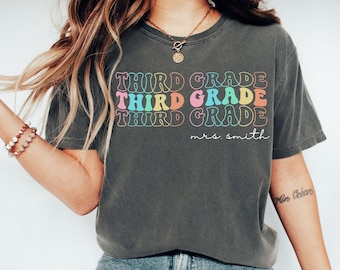 May include: A dark grey t-shirt with the text "THIRD GRADE" repeated three times in rainbow colours. The text "mrs. smith" is written below the repeated text.