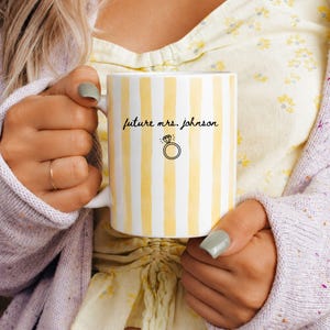 May include: A white ceramic mug featuring vertical yellow stripes and the black text "future mrs. johnson" written in a cursive script above a simple black line drawing of a diamond engagement ring centered on the front of the mug.