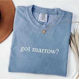 May include: A blue Comfort Colors t-shirt with the phrase "got marrow?" in white. A gold necklace with a pendant is draped across the shirt. A brown hat and dried plant are in the background.