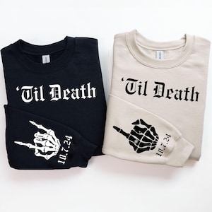 May include: Two sweatshirts, one black and one beige, with the phrase "'Til Death" printed in black gothic font. The black sweatshirt has a white skeleton hand with a ring on the finger making a rock and roll hand gesture on the sleeve. The beige sweatshirt has a white skeleton hand with a ring on the finger pointing on the sleeve. Both sleeves have the date "10.7.24" printed in white.