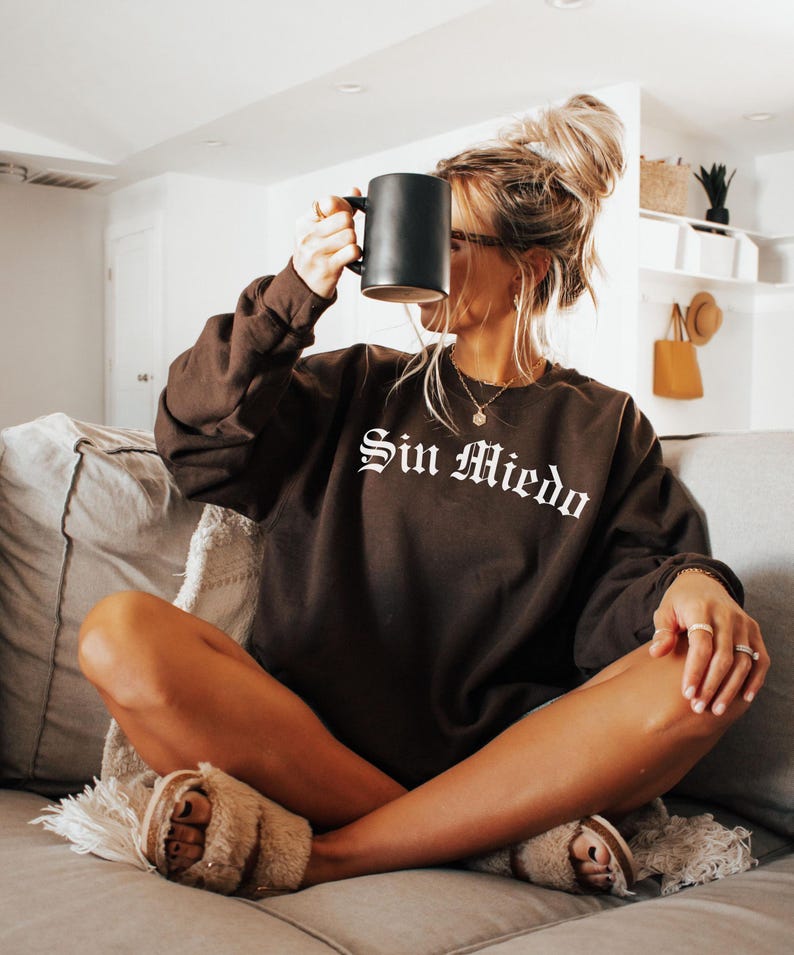 K&ouml;nnte beinhalten: Eine Frau tr&auml;gt ein braunes Sweatshirt mit dem Schriftzug "Sin Miedo" in wei&szlig;en Buchstaben. Sie sitzt auf einem Sofa und h&auml;lt eine schwarze Tasse.