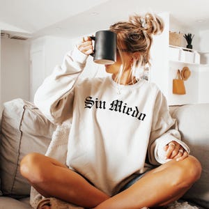 K&ouml;nnte beinhalten: Eine Frau tr&auml;gt ein cremefarbenes Sweatshirt mit dem Text "Sin Miedo" in schwarzen gotischen Buchstaben. Sie sitzt auf einem Sofa mit gekreuzten Beinen und h&auml;lt eine schwarze Tasse.