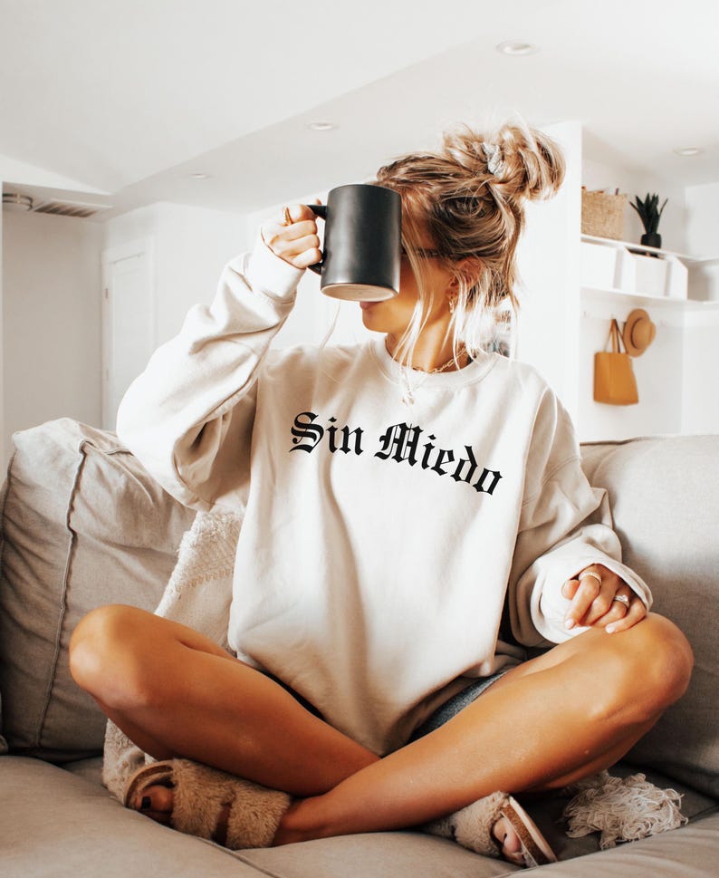 K&ouml;nnte beinhalten: Eine Frau tr&auml;gt ein cremefarbenes Sweatshirt mit dem Text "Sin Miedo" in schwarzen gotischen Buchstaben. Sie sitzt auf einem Sofa mit gekreuzten Beinen und h&auml;lt eine schwarze Tasse.