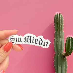 May include: A white sticker with the words "Sin Miedo" in a gothic font is held in front of a pink background. A green cactus with pink spines is to the right of the sticker.