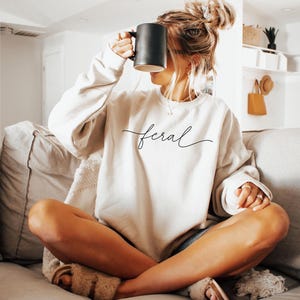 Puede incluir: Una mujer con una sudadera blanca con la palabra "feral" escrita en cursiva en la parte delantera. Est&aacute; sentada en un sof&aacute; con las piernas cruzadas y sosteniendo una taza negra.