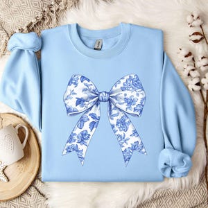 May include: A light blue sweatshirt with a large white and blue floral bow design on the front.