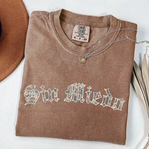May include: A brown short-sleeve t-shirt with a crew neckline and the text Sin Miedo printed in a gothic font with a marbled pattern. The shirt features a Comfort Colors brand label on the inside collar and is paired with a silver chain necklace.