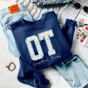 May include: A navy blue sweatshirt with a white and yellow floral print "OT" design. The text "Occupational Therapy" is printed below the design.