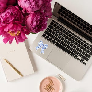 May include: A white laptop computer with a blue and white patterned bow sticker on the top left corner of the trackpad. The laptop is on a white surface with a pink peony bouquet, a white notebook, and a pink dish with gold paper clips.