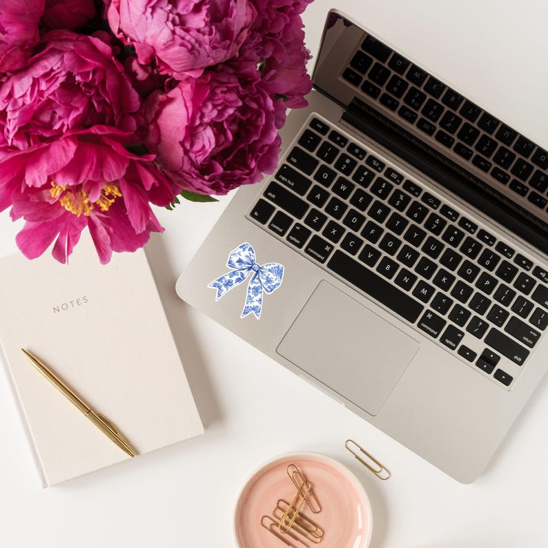 May include: A white laptop computer with a blue and white patterned bow sticker on the top left corner of the trackpad. The laptop is on a white surface with a pink peony bouquet, a white notebook, and a pink dish with gold paper clips.
