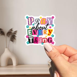 May include: A colorful sticker with the words "PRAY about EVERY thing" in a collage style. The letters are in various colors and fonts, decorated with small flowers. The sticker is held by a person's hand.