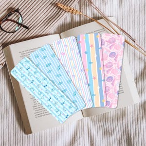 May include: A collection of colorful bookmarks with various patterns, including stripes, seashells, and sunglasses. The bookmarks are displayed on an open book, with a pair of glasses and a sprig of wheat in the background. The bookmarks are made of paper.