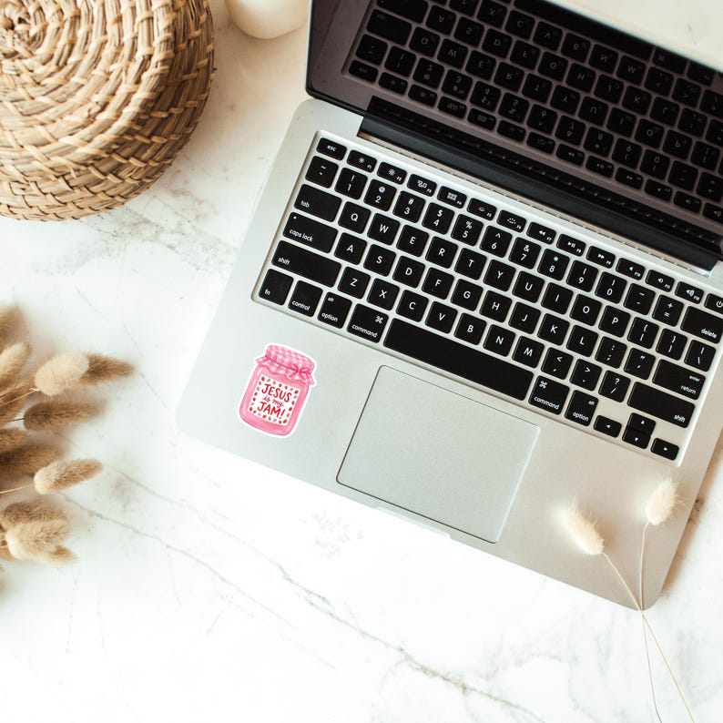 May include: A pink sticker with the text "JESUS JAM" in a jar design is placed on a laptop. The laptop is open on a white marble surface, with a woven basket and dried plants in the background.