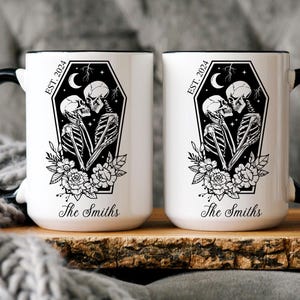 May include: Two white ceramic mugs with black handles and rims. Each mug features a black and white illustration of two skeletons kissing inside a coffin with flowers and the text "EST. 2024" and "The Smiths".
