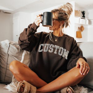 May include: A brown sweatshirt with the word "CUSTOM" in a camouflage-style font. The sweatshirt is worn by someone holding a black mug. The person is sitting on a couch.