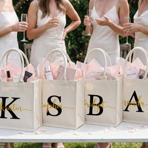 Pode incluir: Quatro sacolas de lona na cor off-white com duas alças, cada uma com uma letra preta grande com serifa e um nome em cursivo dourado na frente. As sacolas estão preenchidas com papel de seda rosa e vários pequenos itens de presente.