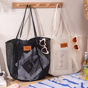 May include: Two mesh tote bags, one black and one off-white, with leather name tags and heart-shaped sunglasses. The black bag has "Stacey" on the tag, and the off-white bag has "Jocelyn". A straw hat and drinks are in the background.